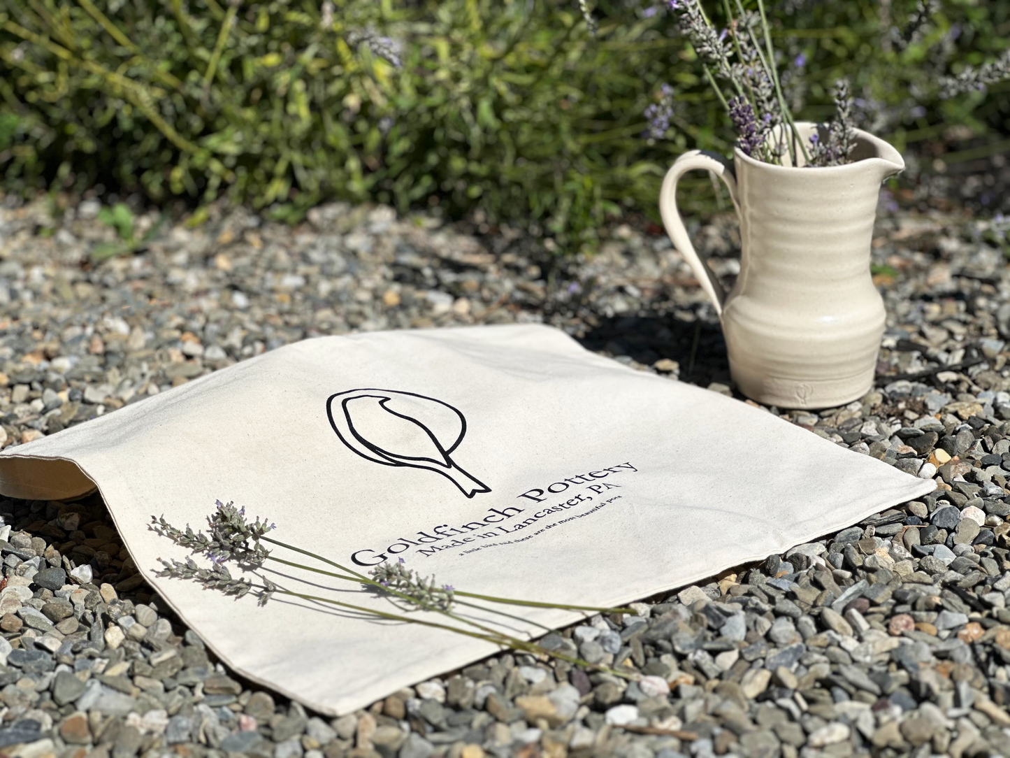 Canvas bag laying on stone gravel next to medium pitcher with lavendar in the pitcher and on the bag outside