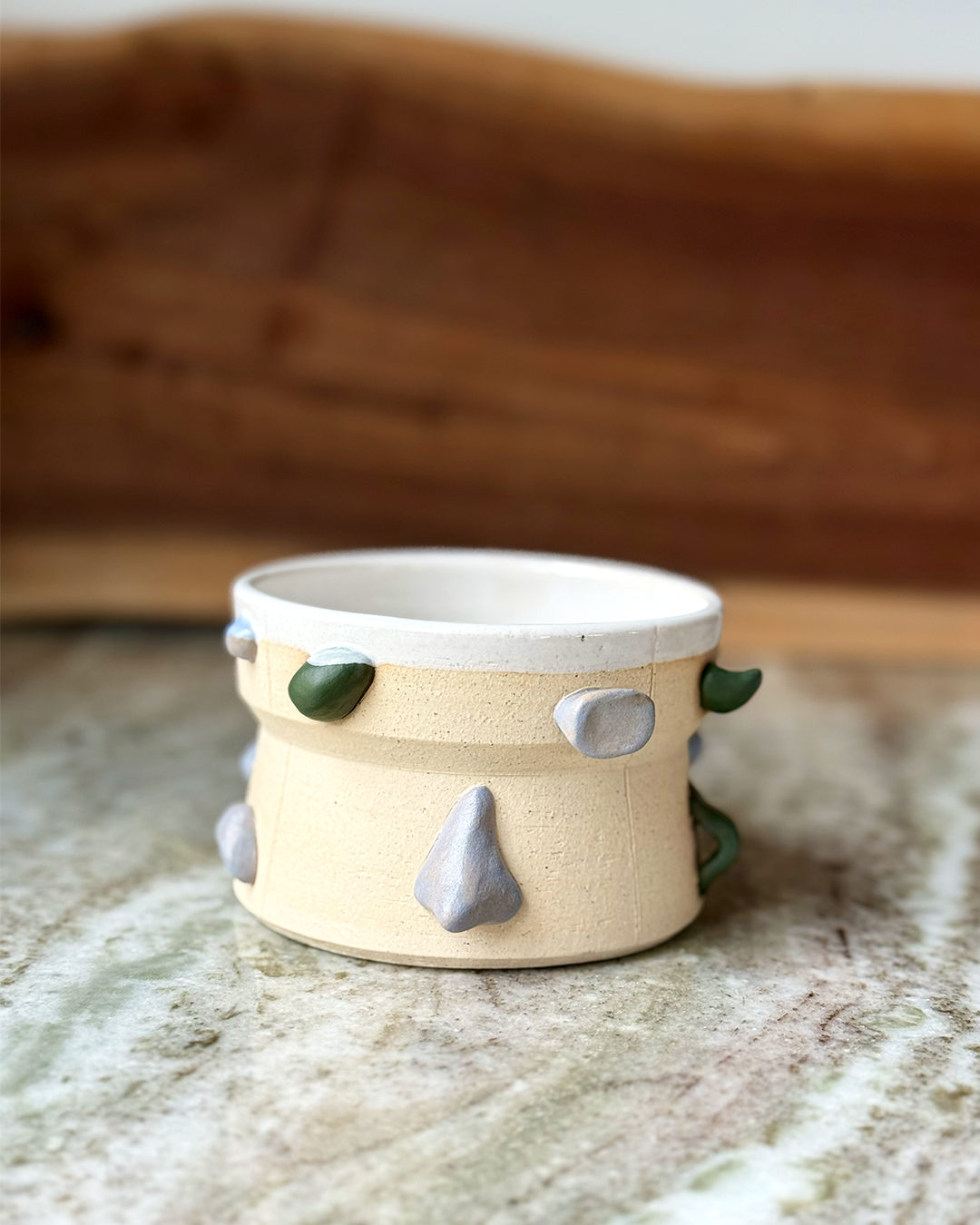 Climbing wall inspired mug with a backdrop of marble and wood. Handholds are green and gray with a white glaze inside on a tan clay body