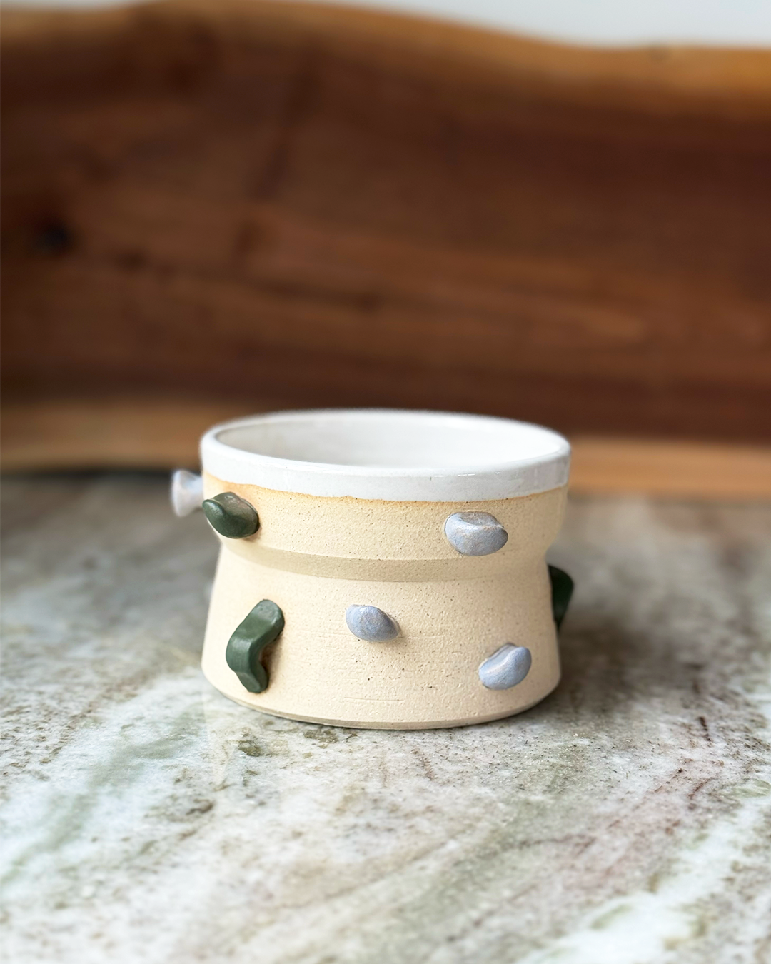 Climbing wall inspired mug with green and gray handholds on a tan clay body with a white glaze inside on a marble counter with a wood backdrop