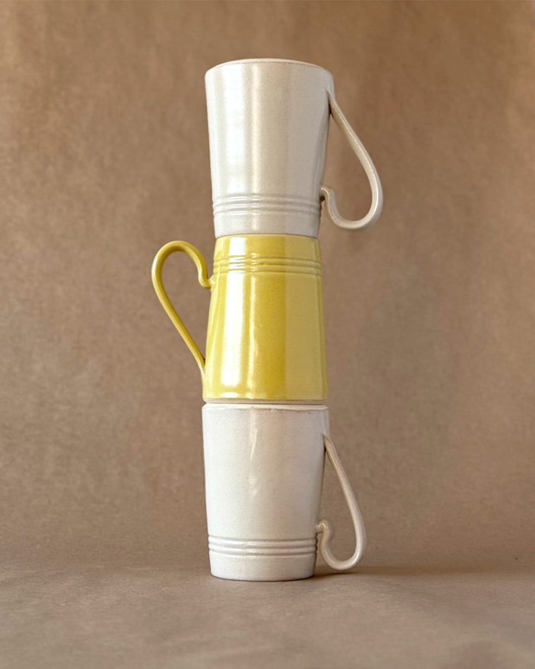 Three Mugs stacked on top of each other. Bottom and Top mug in Linen white glaze, middle in a yellow glaze. Handles facing opposite directions out to the side.