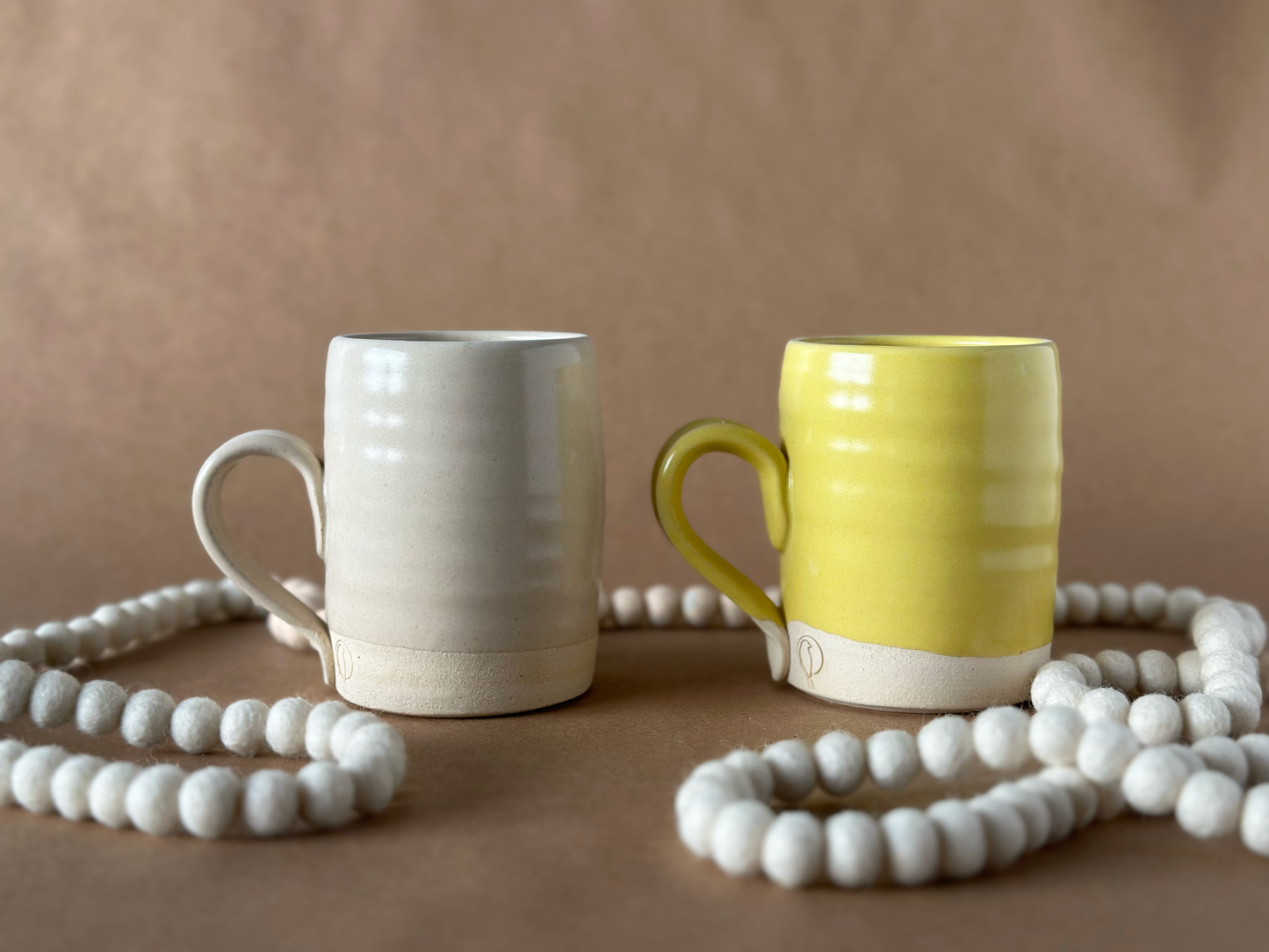 Two Mugs one Linen and One Yellow with a snow needle felted wool garland that is white on a brown backdrop
