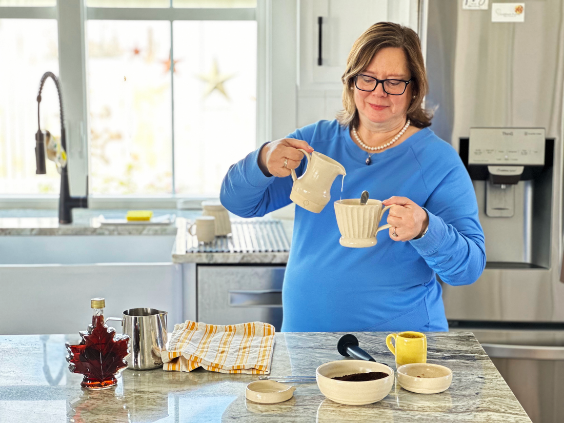 A Woman with brown hair and a blue shirt pouring milk into a handmade pottery mug in a kitchen 