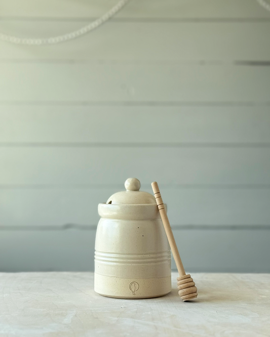 White ceramic honey jar with a wooden honey dipper on a light surface and light gray background