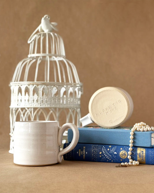 Two Mugs One with the Bottom facing you that says "I Love you More" sitting on a stack of Blue books with a pearl necklace draped to the side. A white birdcage in the back. on a brown backdrop