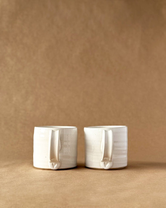 Two White Mugs handles facing you. With Little heart stamps at the bottom of the handle set on a brown backdrop.