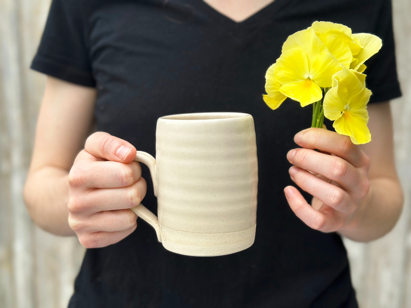 Handmade Ceramic Mug held in one hand. In other hand small bouquet of yellow pansies with a person in a black shirt behind