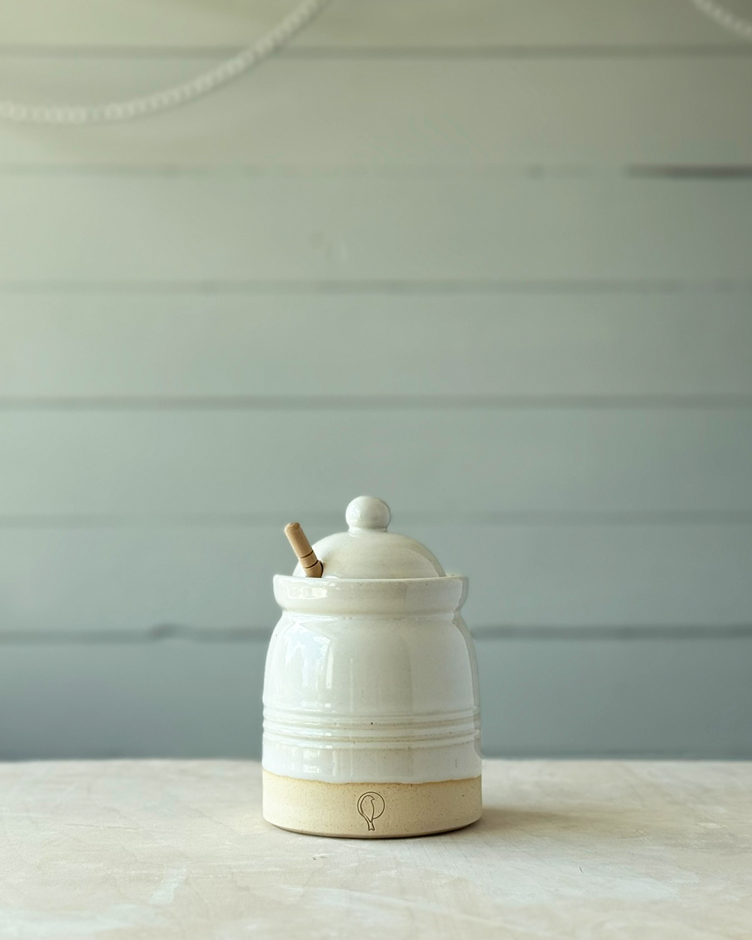 White ceramic jar with wooden handle on a light wooden surface against a light gray wall.