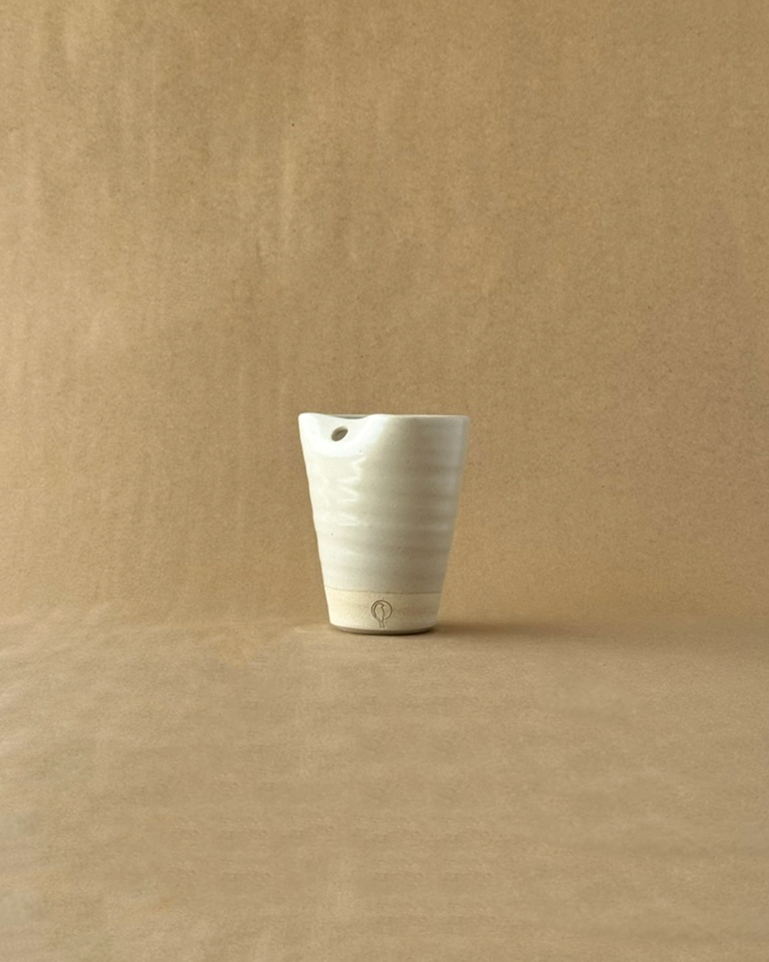 A off-white ceramic cup with a small hole near the rim, standing on a matching beige surface against a textured beige background.