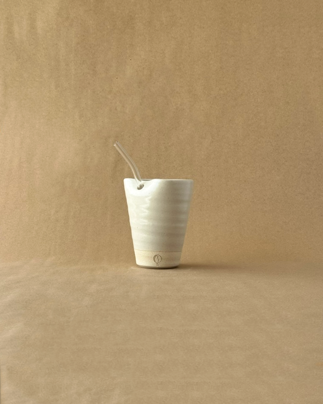 tan tumbler with a glass straw on a brown backdrop with a bird logo